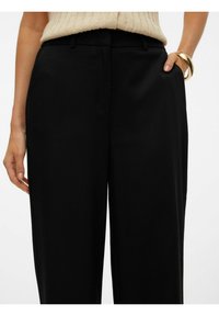 Pantalon large noir avec une texture lisse, taille ajustée et poches latérales, complété par un haut beige clair et un bracelet en or.
