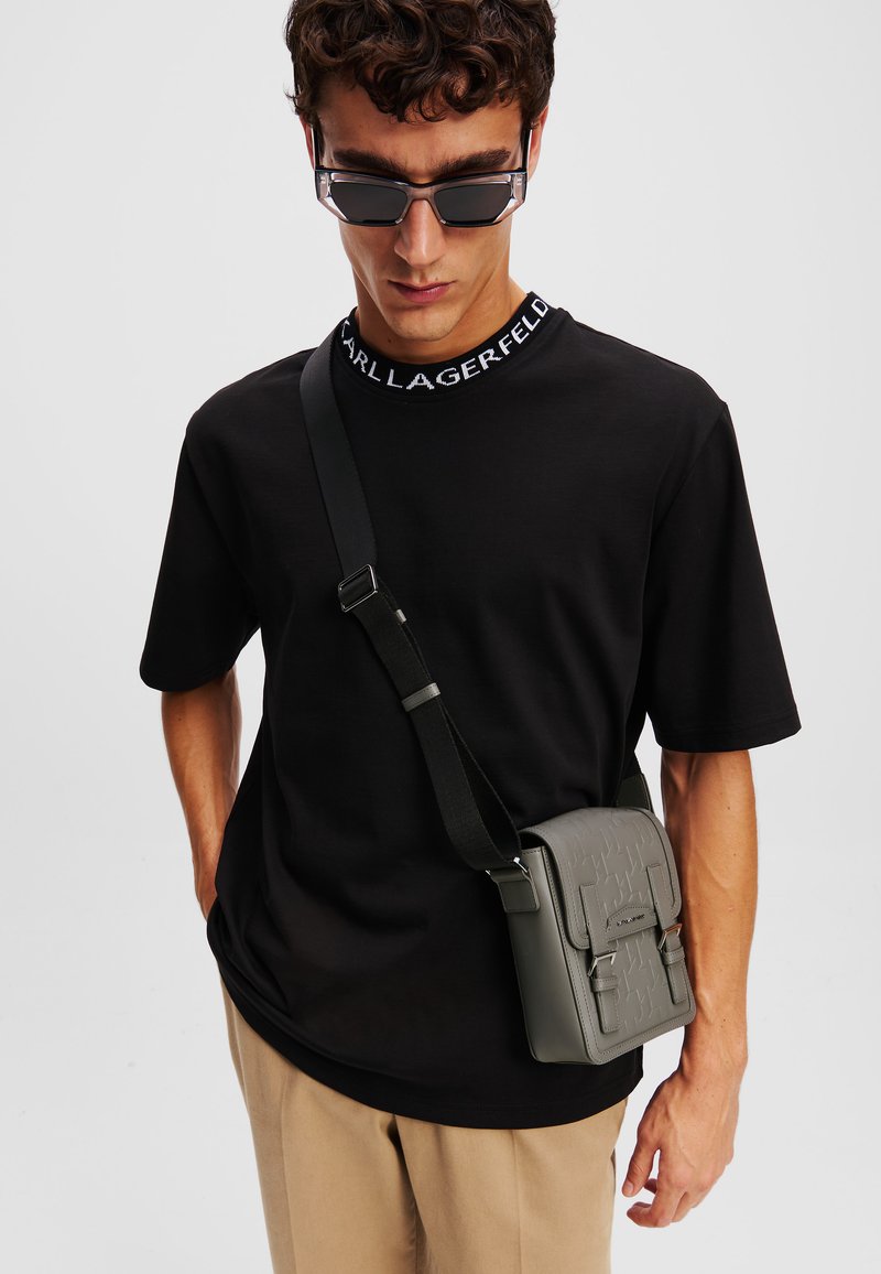 Camicia nera a maniche corte con colletto logo e pantaloni beige. Il modello porta una borsa a tracolla rettangolare grigia con dettagli testurizzati.