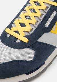 Sneaker con tomaia in rete grigia, accenti in suede blu navy, lacci gialli e suola in gomma testurizzata con dettagli multicolori.