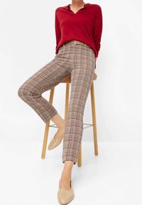 Rode trui met een V-hals, gecombineerd met een geruite broek in beige, bruin en bordeauxrood. Beige instappers ronden de outfit af.