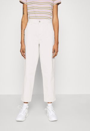 Person trägt weiße, hoch taillierte, gekürzte Hosen, weiße Turnschuhe und ein kurzärmeliges gestreiftes Hemd mit lila, gelben und weißen Linien.