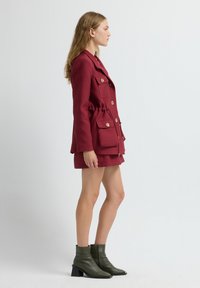 Donna che indossa un blazer borgogna strutturato e una gonna abbinata con grandi bottoni dorati, abbinati a stivaletti con tacco verde scuro, in posa di profilo.