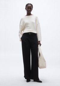Krém színű, túlméretezett pulóver nyers szegéllyel, fekete, bőszárú nadrággal és bézs színű tote táskával párosítva. Sima anyagok és laza szabás.