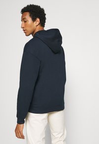 Marineblauer Hoodie mit weicher Textur, verstellbarem Kordelzug und gerippten Bündchen sowie Saum. Getragen mit hellen Hosen, die einen lässigen Sitz zeigen.