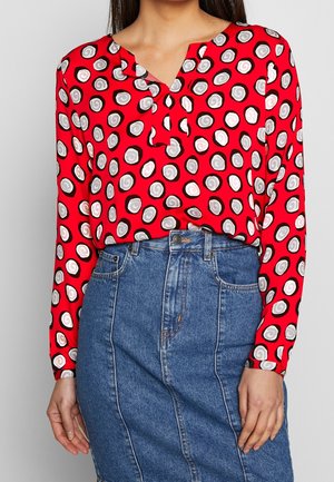 Woman wearing a red long-sleeve blouse with white and gray spiral circles tucked into a high-waisted blue denim skirt.