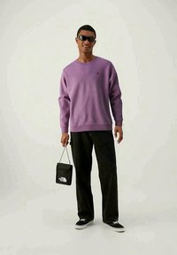 Felpa viola chiaro con polsini a coste, pantaloni scuri, scarpe nere e una piccola borsa a tracolla nera con dettagli del logo. Il modello indossa occhiali da sole.