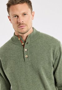 Pull en maille verte avec un col côtelé haut, trois boutons et des accents rouges contrastants près de l'encolure. Texture douce, design décontracté.