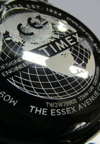 Cadran noir avec un graphisme mondial argenté, featuring la marque et le texte 'TIMEX'. Le design comprend des lignes et des coordonnées, avec un dos en acier inoxydable.