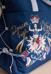 Tamsiai mėlyna audinio rankinė su grafiniu atspaudu, vaizduojančiu moterį ir vyrą, žvaigždes, Jungtinės Karalystės vėliavas ir čiužinėjimo pačiūžas, su šviesiai pilkomis rankenomis.