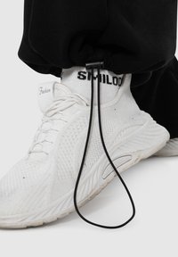 Hvide teksturerede sneakers med snørebånd, parret med sorte sweatpants og en hvid sok med teksten "Fashion" og "STILOD" i sort.