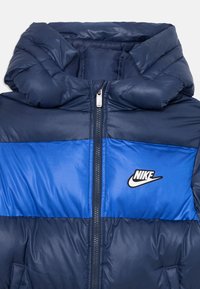 Pufová bunda v tmavomodrej a jasne modrej farbe, s rôzne zipsovým zapínaním a kapucňou. Logo Nike zobrazené na hrudi. Hladká textúra.