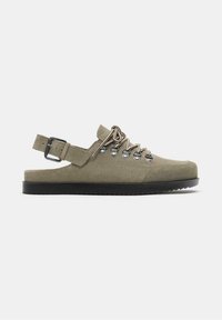 Chaussure beige en daim à dos ouvert avec laçage à l'avant, œillets en métal, bride à boucle réglable, et semelle plate en caoutchouc noir.