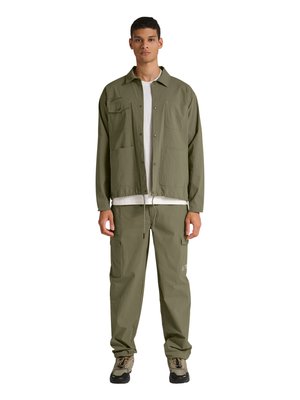 Ung mand iført olivengrøn jakke og cargobukser med beige og sorte vandrestøvler, stående med fronten mod kameraet mod hvid baggrund.