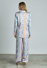 Ensemble blazer et pantalon à motifs dans des couleurs pastel, avec des designs géométriques, une texture lisse et des manches longues. Des talons argentés complètent la tenue.