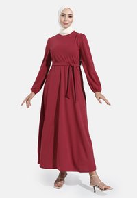 Lange, bordeauxrode jurk met pofmouwen en een riem in de taille. Gemaakt van zachte stof met een vloeiende uitstraling, die net onder de enkels reikt.
