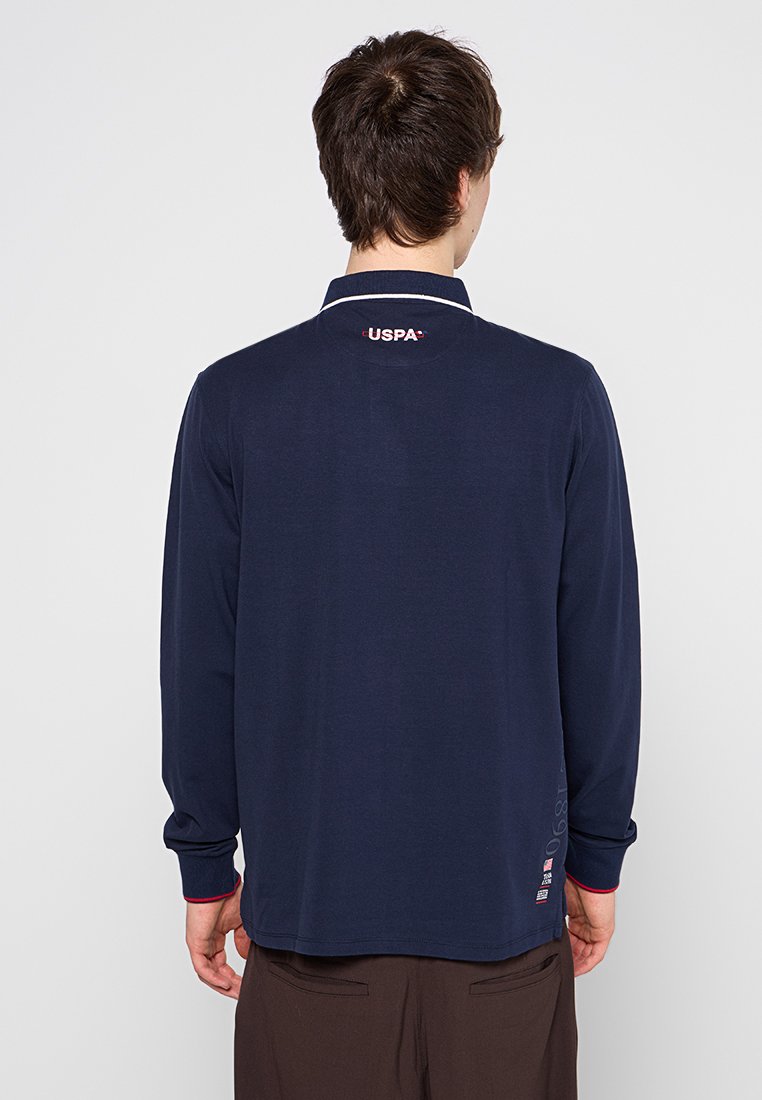 Polo de manga larga de color navy con cuello blanco, que presenta un logotipo rojo en la parte posterior del cuello y detalles rojos en los puños. Tela suave y lisa.