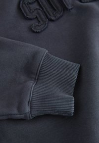 Felpa grigia scura con polsini a coste e un design del logo testurizzato. Tessuto morbido con una finitura liscia e cuciture strutturate.