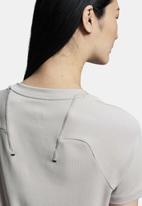Camisa de manga corta gris claro con una textura suave, cuello redondo y costuras decorativas, con cordones en el cuello de la parte trasera.