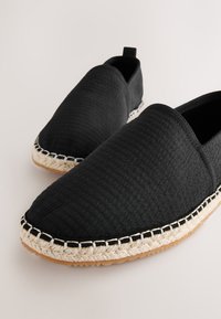 Black slip-on shoes with a textured fabric upper, featuring a jute rope sole and contrasting stitching along the edge.
