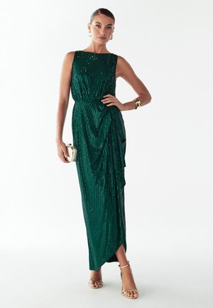 Zielona sukienka maxi wykonana z połyskujących cekinów, z dopasowanym gorsetem, marszczeniami i rozcięciem po boku. Dopełniona złotą biżuterią i kopertówką.