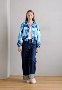 Giacca bomber blu con motivi a onde, abbinata a jeans in denim scuri a vita alta e sneaker bianche. Orecchini rotondi semplici in mostra.