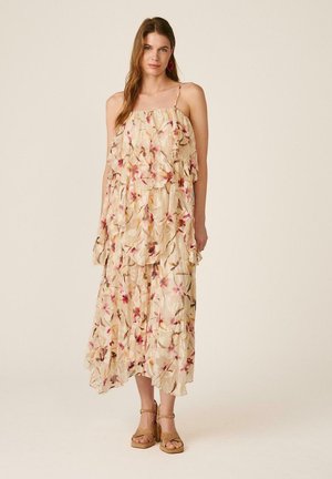 Mujer con un vestido midi beige con flores en capas, tirantes finos y sandalias de tacón ancho color cuero claro, posando contra un fondo claro.