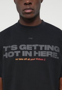 Κοντινό πλάνο ενός ατόμου που φοράει μαύρη μπλούζ�α με το κείμενο "ΓΙΝΕΤΑΙ ΖΕΣΤΟ ΕΔΩ." σε γκρι και "οπότε βγάλε όλα τα ρούχα σου ;)" σε πορτοκαλί και κόκκινο.