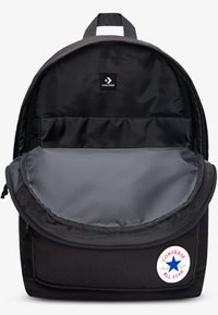 Černý plátěný batoh se šedým vnitřkem. Má zaoblenou klopu, nastavitelné popruhy a kulatý logotyp na přední straně.