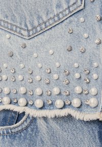 Jeansjacka med ljusblå tvätt, prydd med pärlor och strass. Omlottkanter med slitna, fransiga kanter.