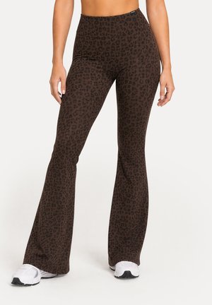 Femme portant des leggings évasés imprimé léopard marron et des baskets blanches, vus de la taille aux pieds sur un fond uni.