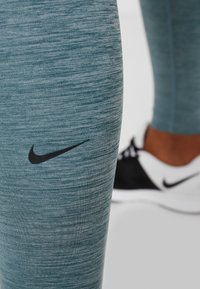 Türkise Fitness-Leggings aus einem strukturierten Stoff, mit einem schwarzen Nike-Logo am linken Oberschenkel, kombiniert mit schwarz-weißen Sportschuhen.