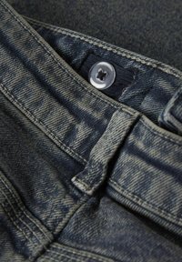 Nærbilde av midjebåndet på mørkeblå denimjeans med sølvfarget knapp og synlige sømmer.