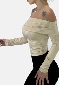 Off-Shoulder, enganierter Oberteil aus leicht beigem, dehnbarem Stoff mit geraffter Textur; lange Ärmel und schlanke Silhouette, kombiniert mit schwarzen Hosen.