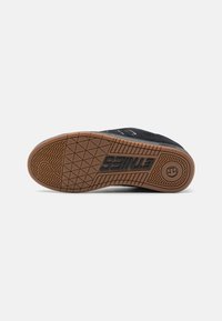 Black suede sneaker with a rubber sole, featuring a textured gum outsole and embossed logo on the heel. Flat laces and stitched accents.