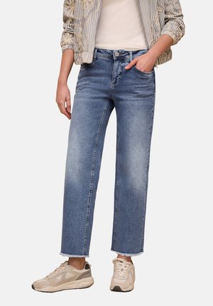 Persoon die lichtgewassen cropped rechte jeans met rafelige zoom draagt, een gestreept kanten jasje, een wit topje en beige sneakers, staand met één hand in de zak.