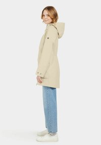 Veste à capuche beige avec une taille à cordon, fermeture éclair à l'avant, poignets élastiques et poches latérales, associée à un jean bleu clair et des chaussures blanches.