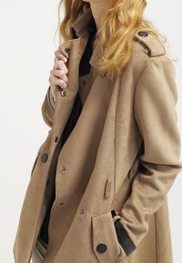 Kobieta w beżowym podwójnym płaszczu z czarnymi guzikami i naramiennikami, ręka dostosowująca kołnierz, z luźnymi blond włosami widocznymi.