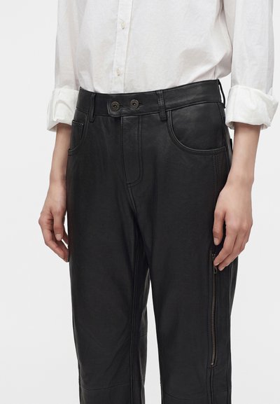 Personne portant un pantalon en cuir noir avec des poches avant et des fermetures éclair, associé à une chemise blanche boutonnée avec les manches retroussées.