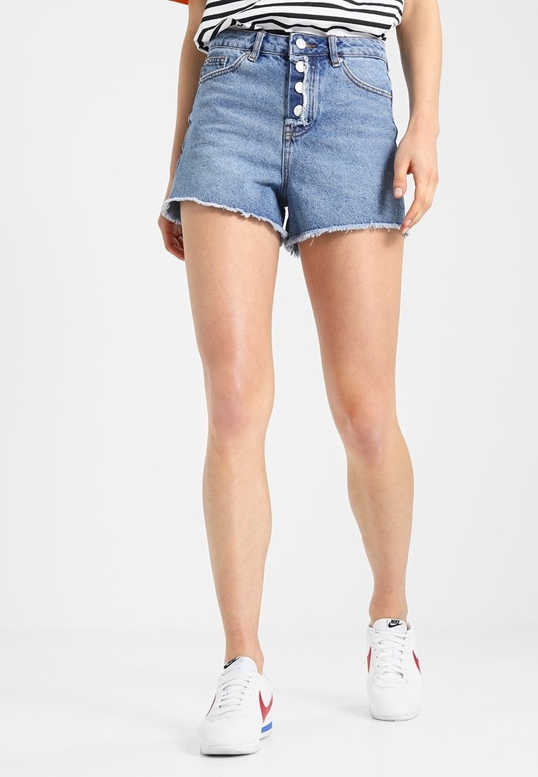Personne portant un short en denim bleu taille haute avec braguette à boutons et des baskets blanches avec des détails rouges et bleus marchant sur un fond uni.