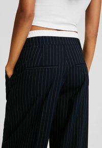Marineblauwe pinstriped broek met een elastische tailleband, voorzien van twee zijzakken en een strak ontwerp. De stof lijkt glad en op maat gemaakt.