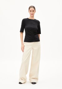 Schwarzes Kurzarmoberteil mit geraffter Textur an der Vorderseite, kombiniert mit hochtaillierten, weiten cremefarbenen Hosen und schwarzem Schuhwerk.