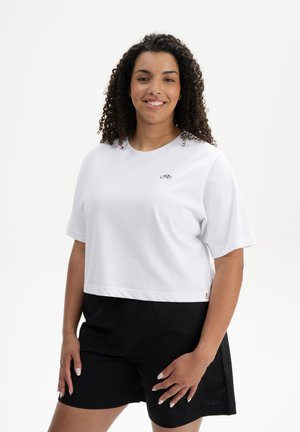 Weißes, kurzes Baumwoll-T-Shirt mit kurzen Ärmeln und einem kleinen schwarzen Fahrrad-Logo auf der Brust, kombiniert mit schwarzen Shorts.