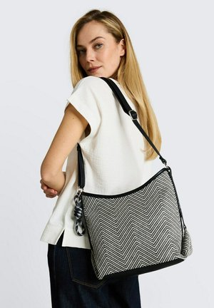 Blondynka w białej bluzce i ciemnych dżinsach niesie dużą czarno-białą torbę na ramię w wzór jodełki z ozdobnym plecionym paskiem.