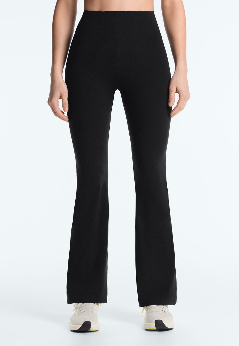 Femme portant un pantalon noir évasé taille haute et des baskets blanches, debout avec les mains détendues le long du corps sur un fond clair.