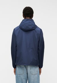 Άνδρας που φοράει ναυτικό μπλε μπουφάν με κουκούλα και ανοιχτόχρωμο τζιν, στέκεται γυρνώντας την πλάτη σε φωτεινό απλό φόντο.