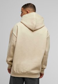 Beige overdimensjonert hettegenser med stor hette, laget av mykt stoff. Har senkede skuldre og ribbet kant. Glatt tekstur, minimal detaljering.
