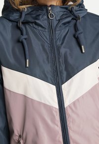 Giacca leggera color-block navy e rosa con cappuccio, zip e cordini regolabili. Texture liscia e dettagli a zigzag.