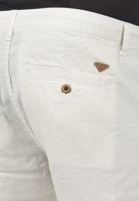 Pantalones de algodón blancos con una etiqueta de marca marrón en forma de triángulo, un único bolsillo trasero con un botón de madera y un acabado de tela texturizado.