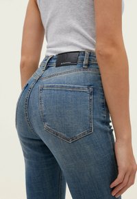 Jeans de denim azul claro con un diseño ajustado, estilo de cinco bolsillos y una etiqueta negra en la cintura trasera. Textura desgastada en la tela.