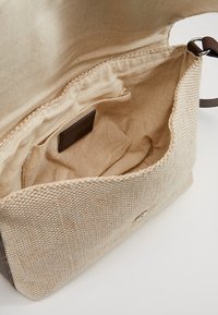 Beige vävd tygaxelväska med öppen klaff, som visar mjukt innerfoder, innerficka och brunt läderrem med metallring.